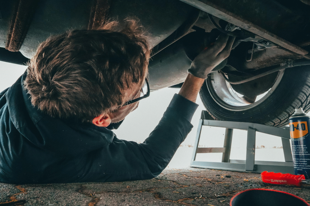 A man is crouched under a car, engaged in maintenance work beneath the wheels.