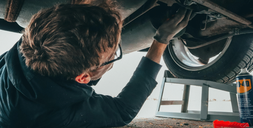 A man is crouched under a car, engaged in maintenance work beneath the wheels.